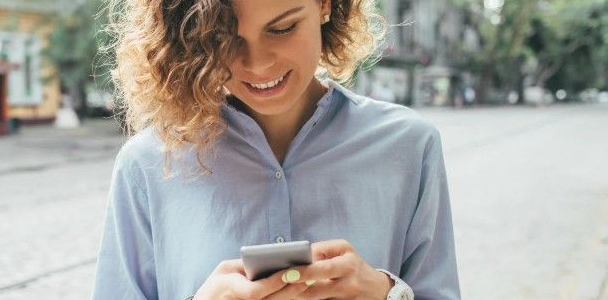 Woman with curly hair smiles while looking at her phone outdoors.