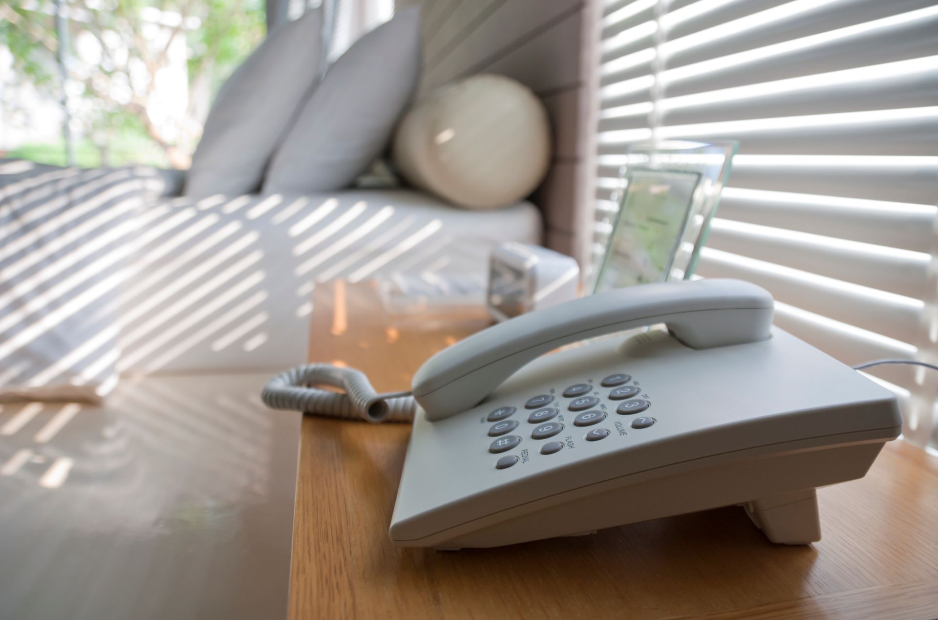 White landline phone on a wooden nightstand next to a window with slatted blinds and a window seat with pillows.