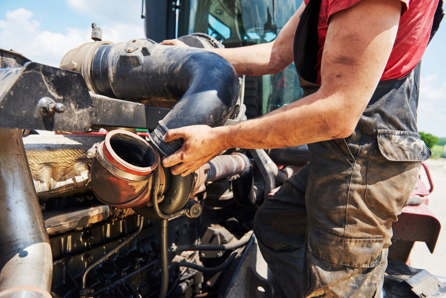 Mechanic working on the engine of a tractor, wearing overalls and a red shirt. Outdoors, sunny day.