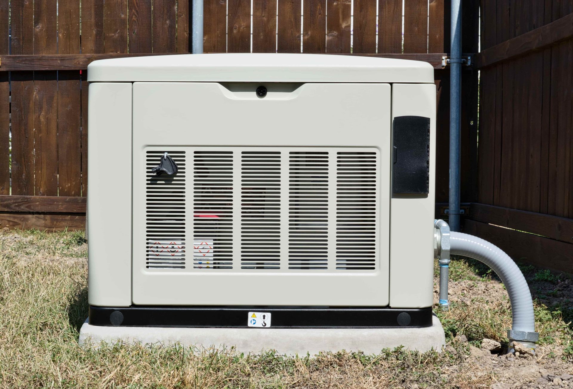 Beige home generator next to a brown wooden fence, on a concrete pad and grass.