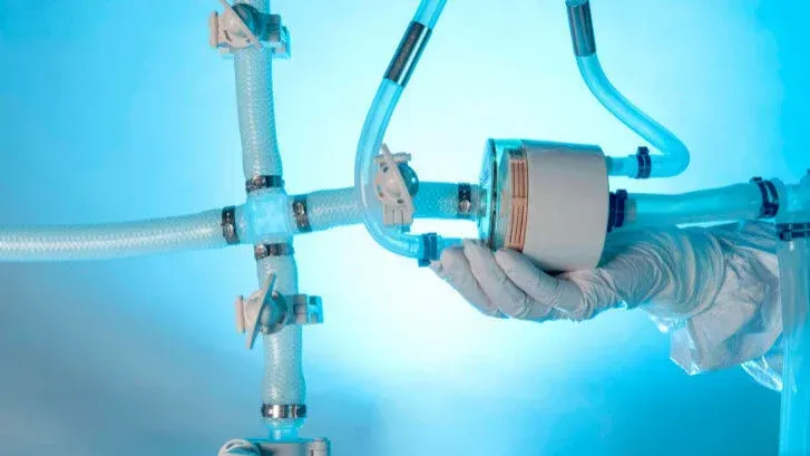 A gloved hand holds a white cylindrical device connected to translucent tubes. Blue background.