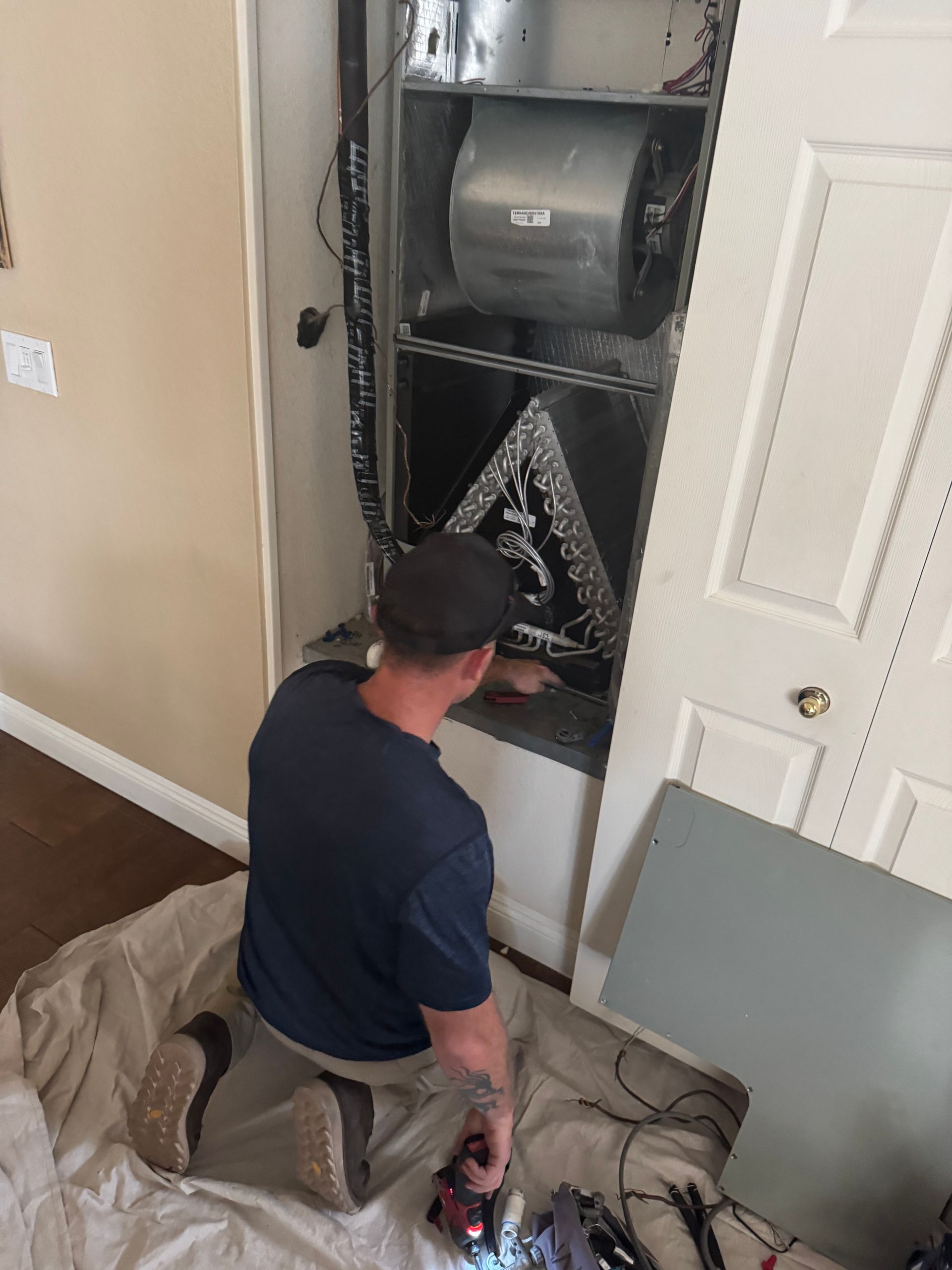 HVAC technician working on an air handler unit inside a closet, kneeling with tools, light interior.