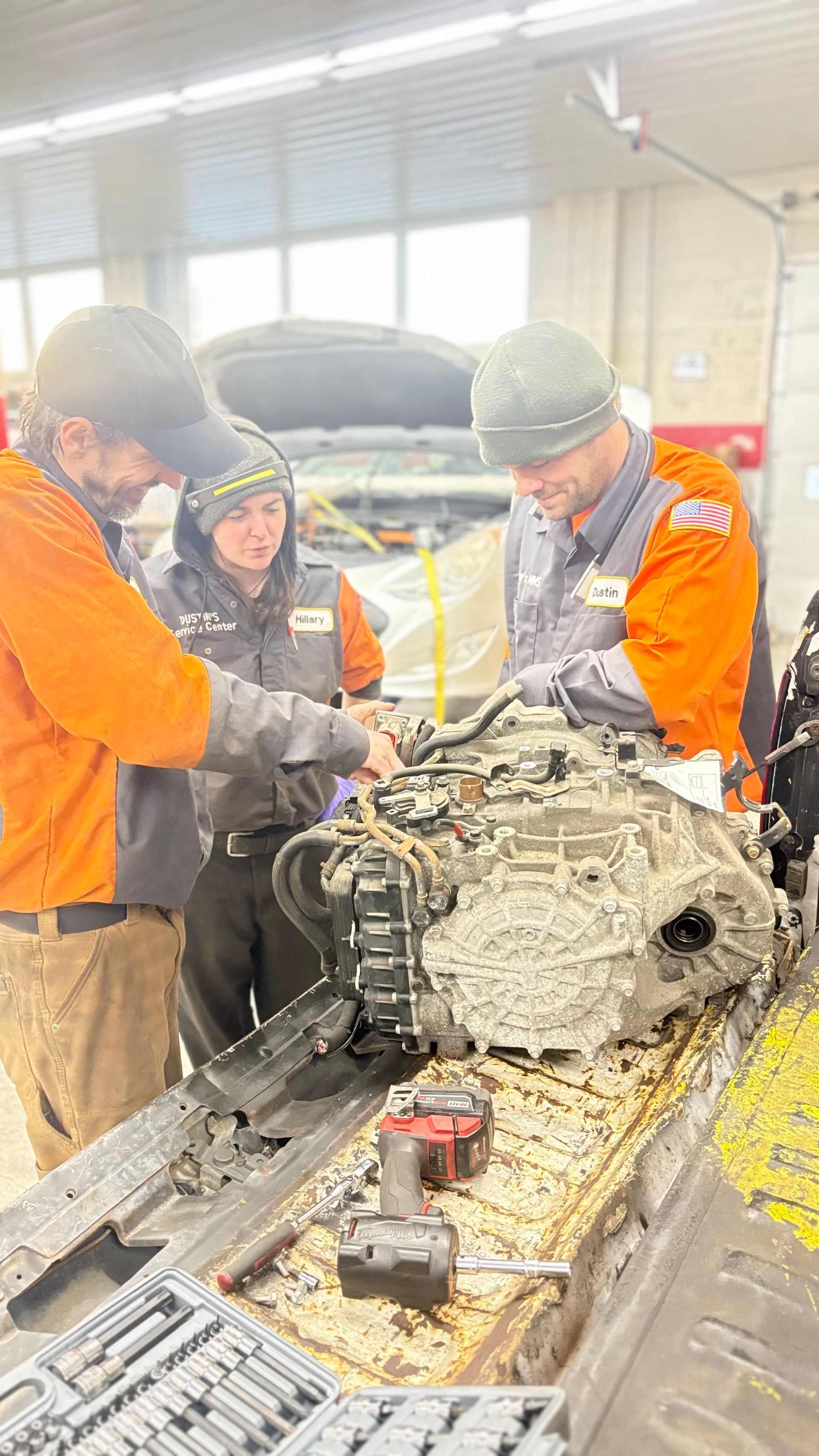 Three mechanics examine engine parts on a workbench in a shop. They wear orange jackets, focusing intently. | Dustin's Automotive Service Center