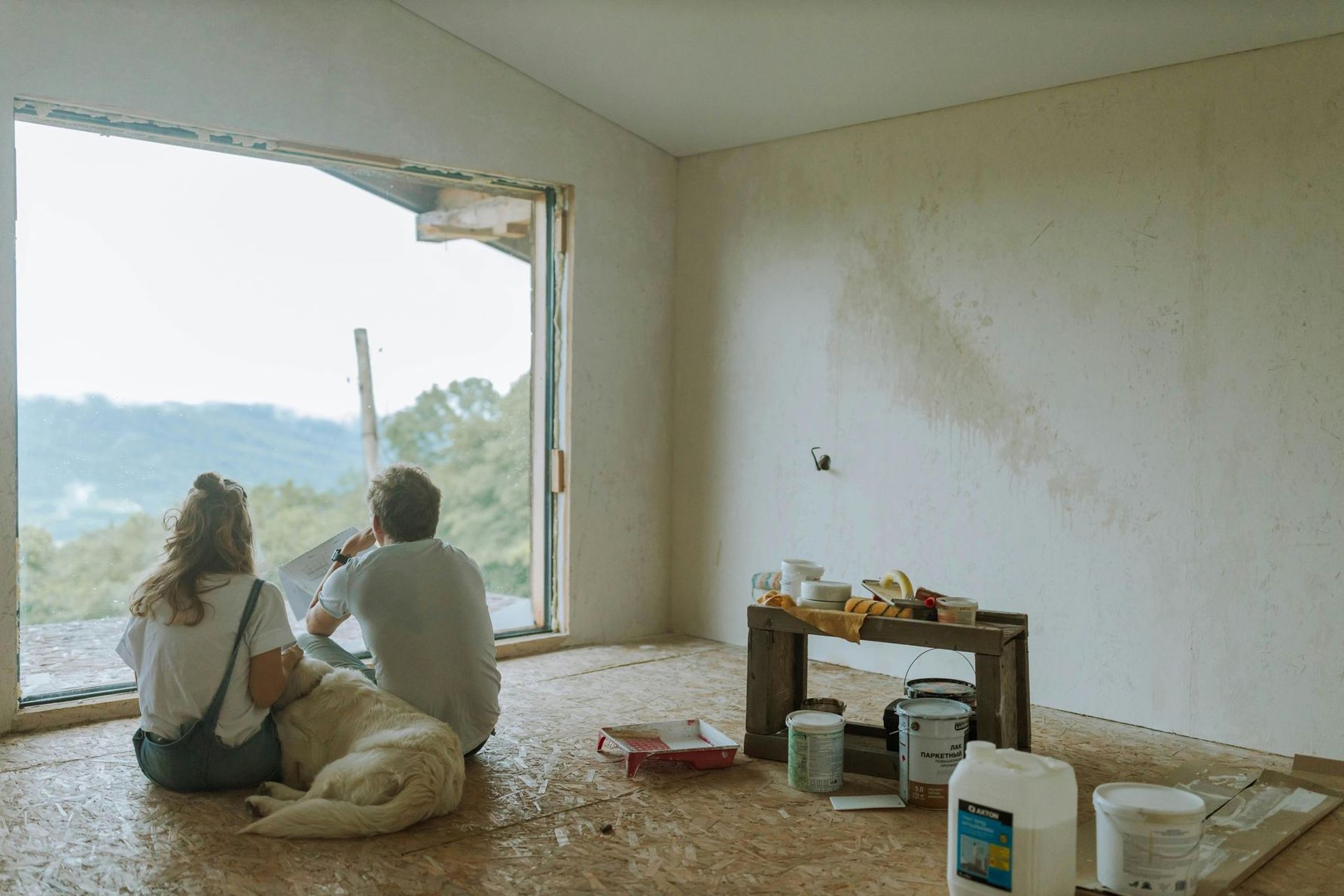 A man and a woman are sitting on the floor with a dog looking out of a window.