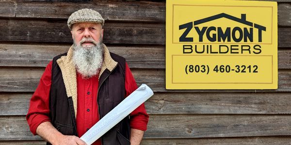 A man with a beard is standing in front of a sign for zygmont builders.