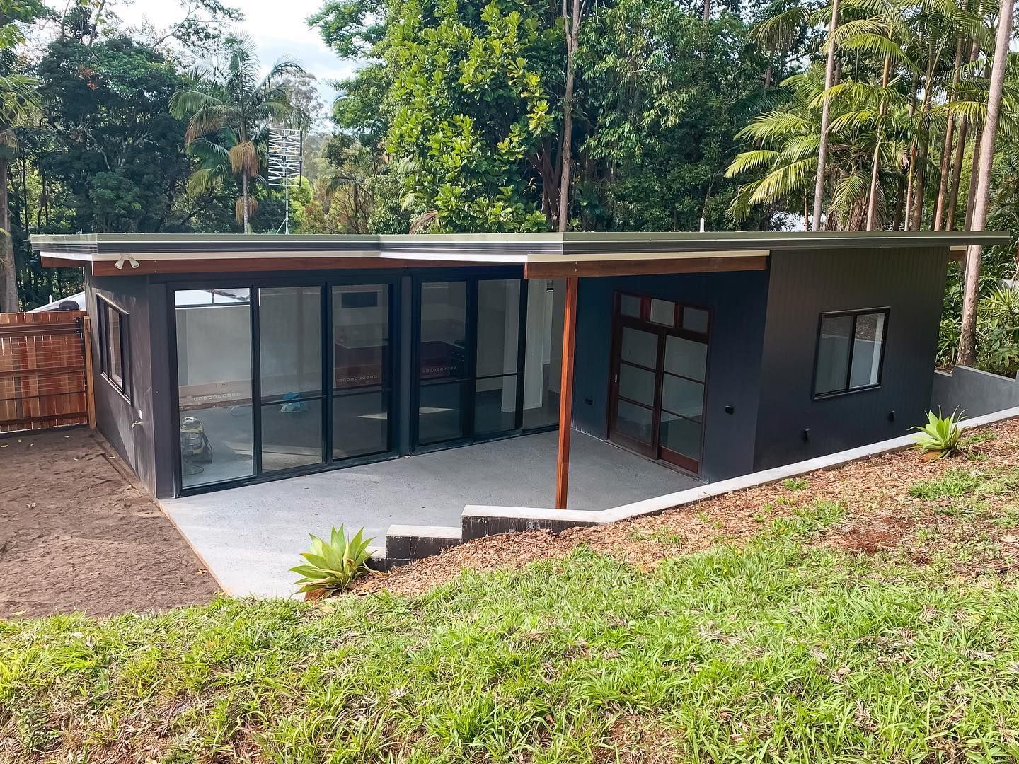 Modern, dark grey building with glass walls and a concrete patio, set in a grassy area surrounded by trees.