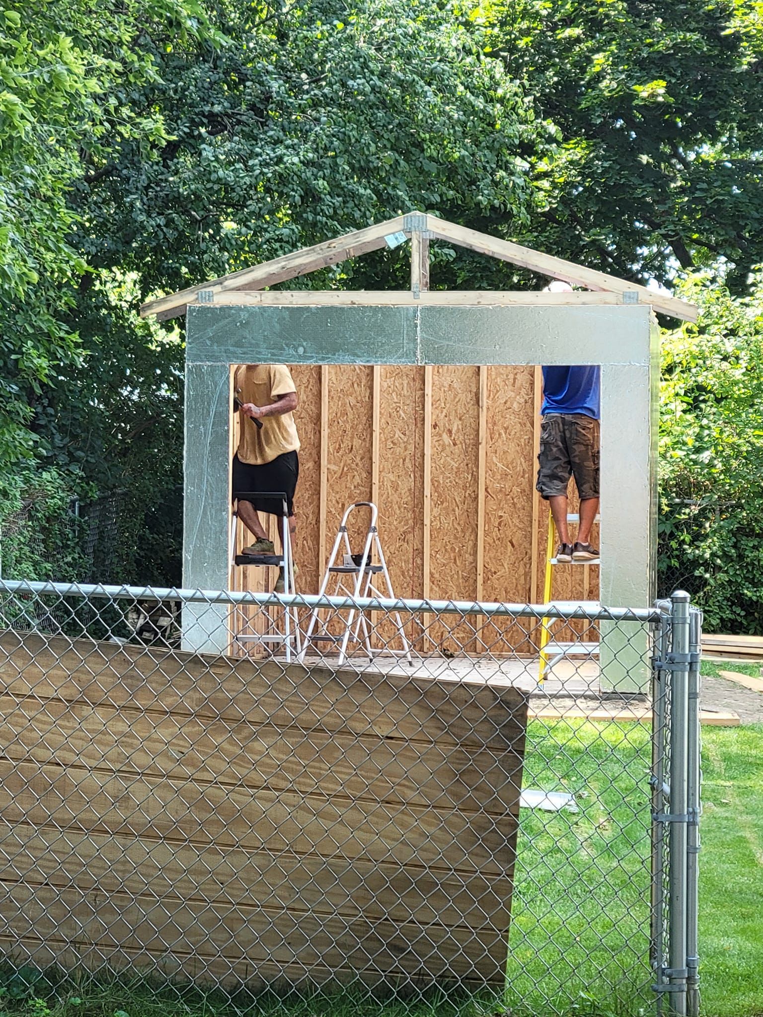Two men are working on a shed in a backyard behind a chain link fence.