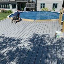 A man is kneeling on a wooden deck next to a swimming pool.