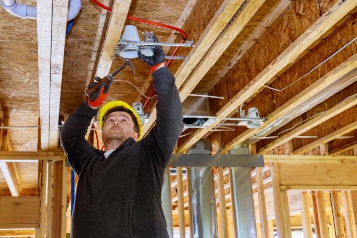Electrician in a yellow hard hat installing wiring in a wooden ceiling.