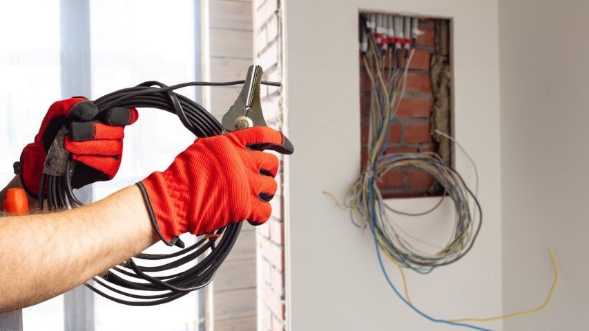 Person wearing red gloves using wire strippers on a black cable near an open electrical box.