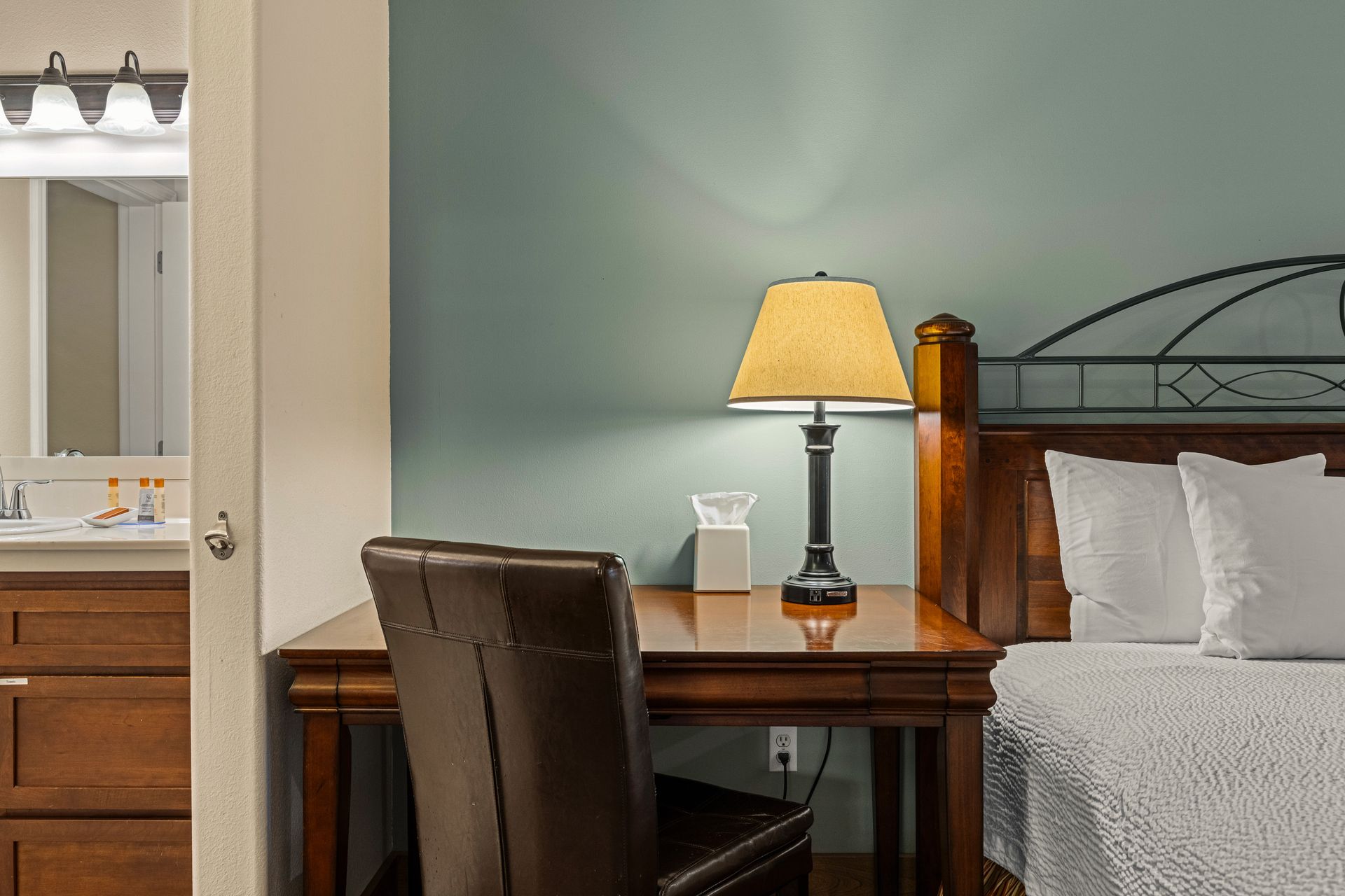 Bedroom corner with desk, chair, lit lamp, and bed beside a bathroom vanity