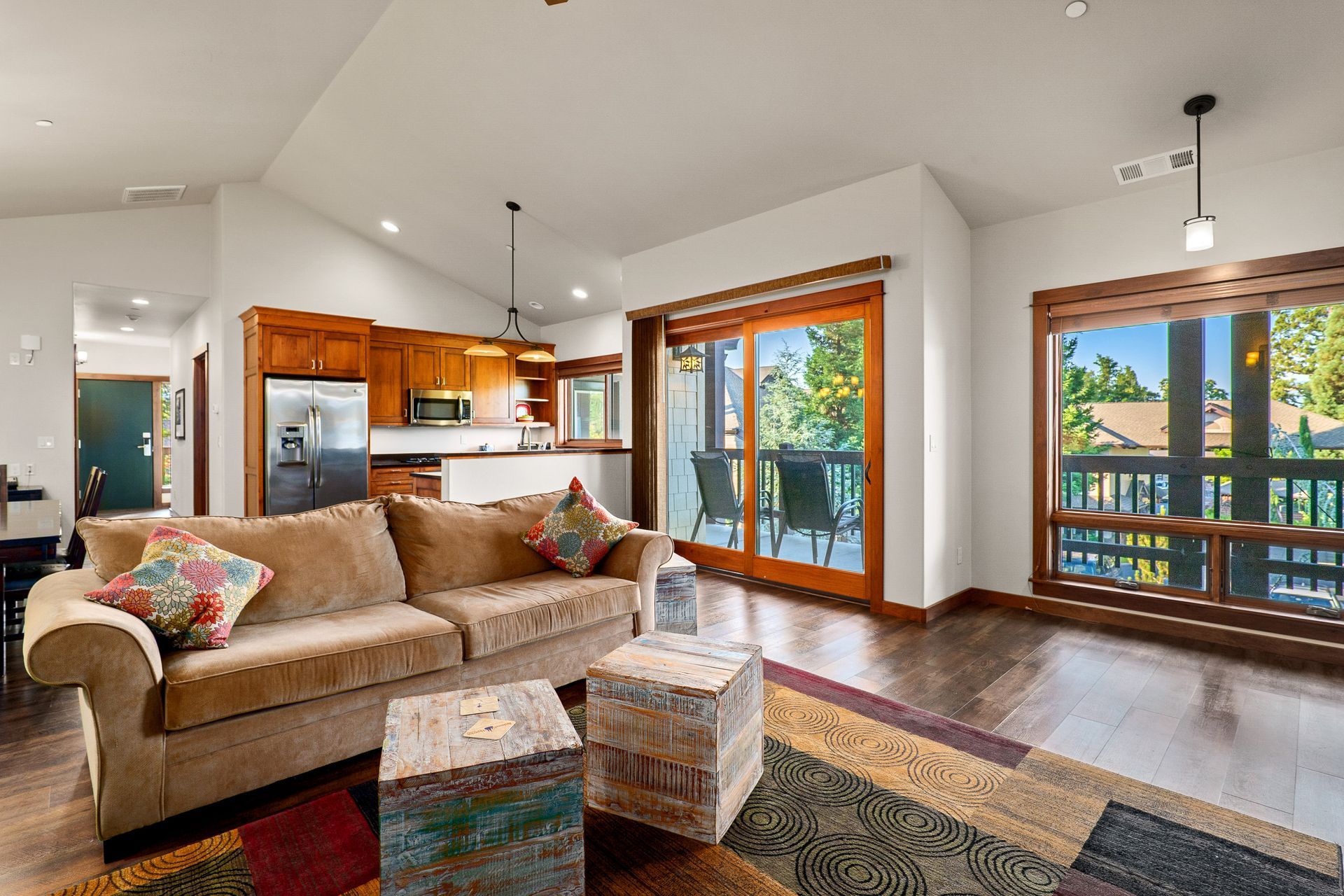 Open living room with beige sofa, colorful rug, kitchen, and large windows overlooking a deck.