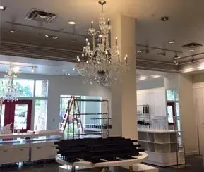 A brightly lit retail interior featuring a large ornate crystal chandelier above a display table with black tiered shelves.