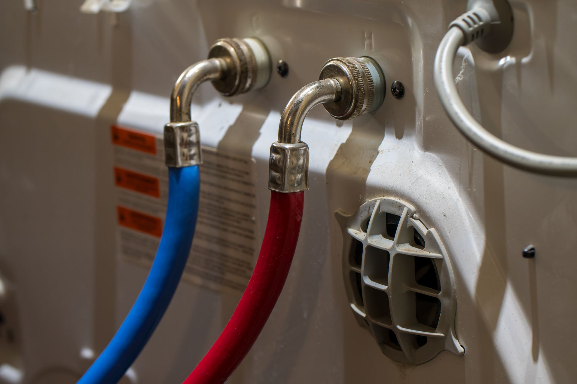 The back of a washing machine with blue and red water hoses connected to the inlets, and a white power cord visible.