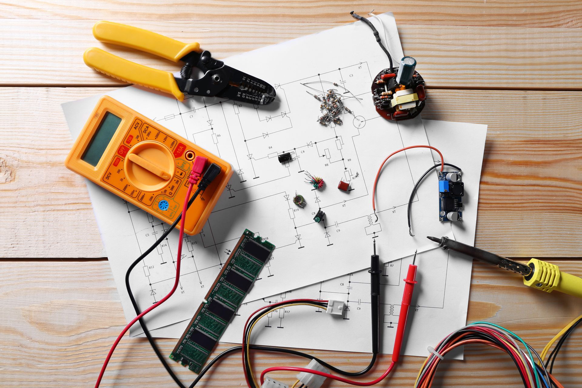 Electronics repair tools and components scattered on a white wooden table, with a multimeter, wires, and pliers.