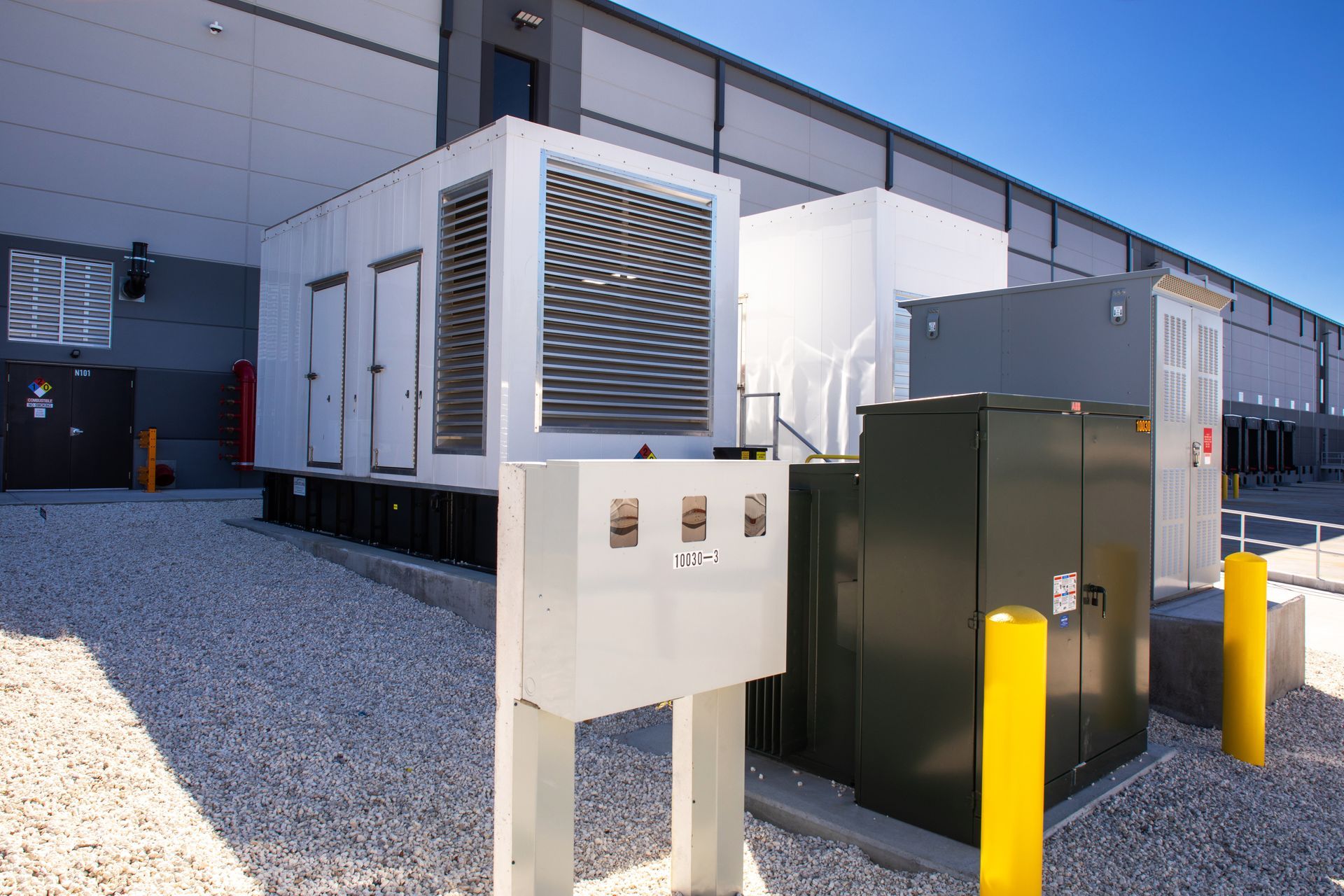 A large industrial generator, electrical cabinets, and safety bollards sit on a gravel lot outside a warehouse building.