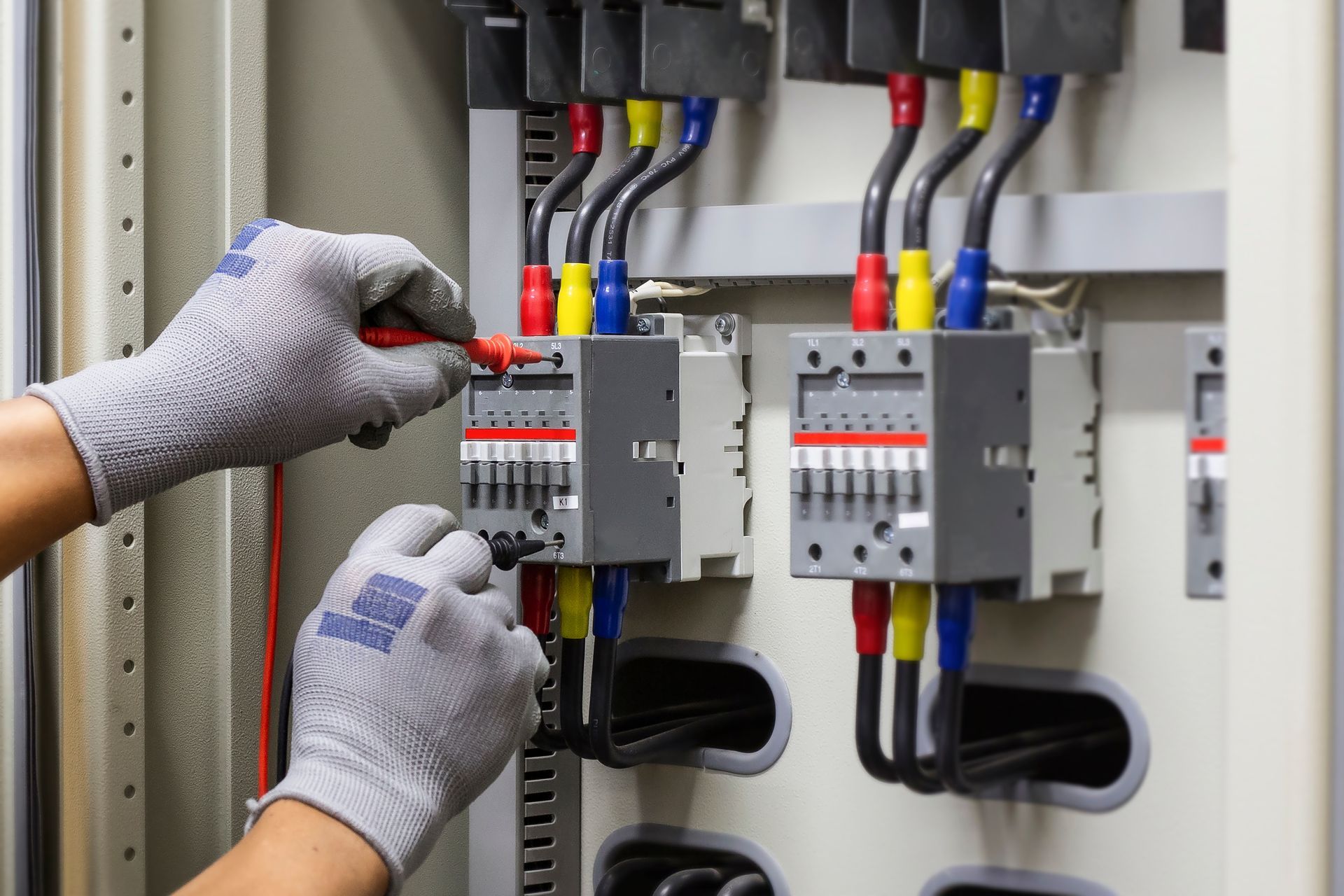 An electrician in grey gloves uses a multimeter to test electrical components inside a control panel.