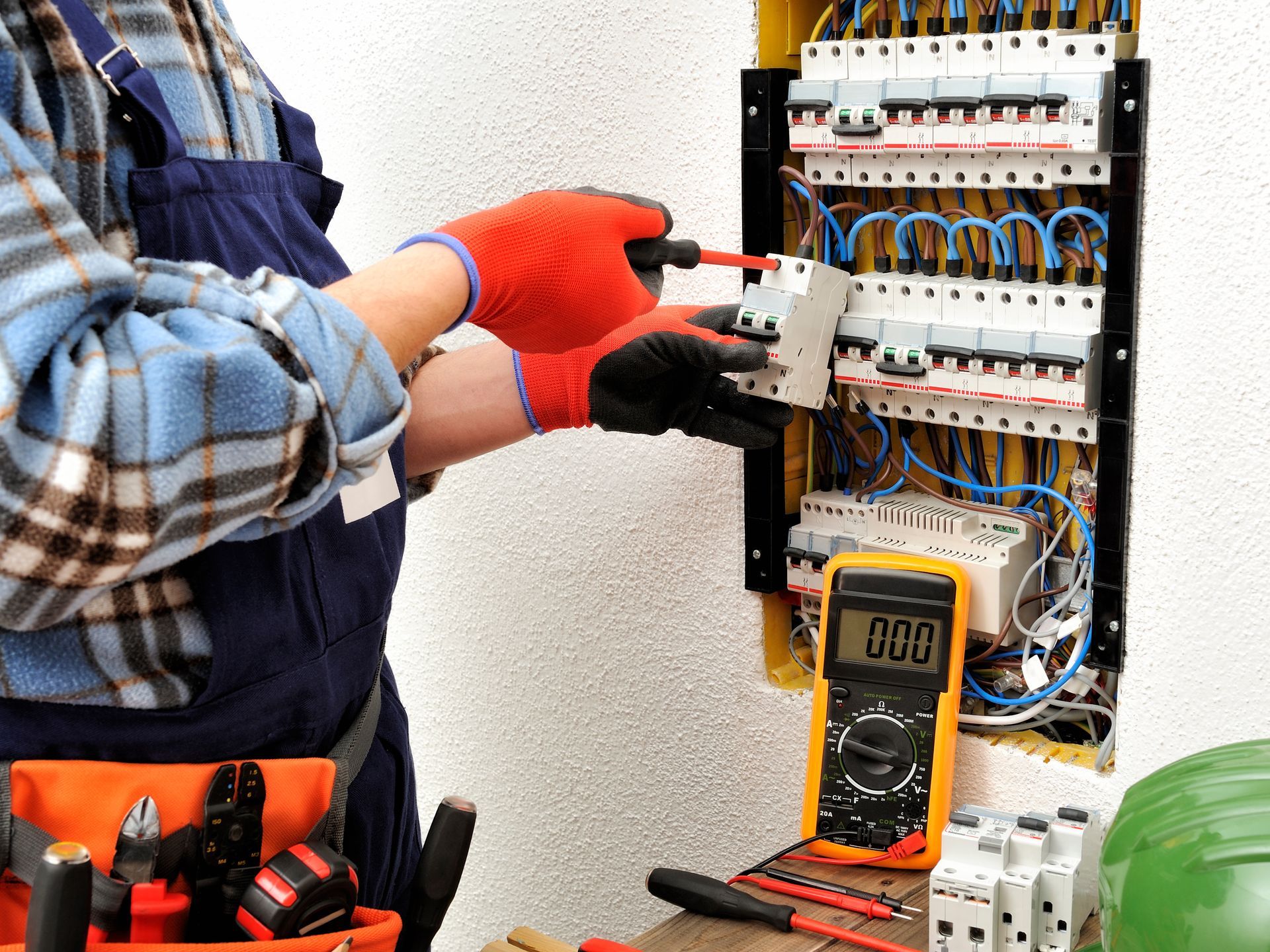 An electrician in workwear installs a breaker in a residential fuse box, using a multimeter to check the electrical circuit.