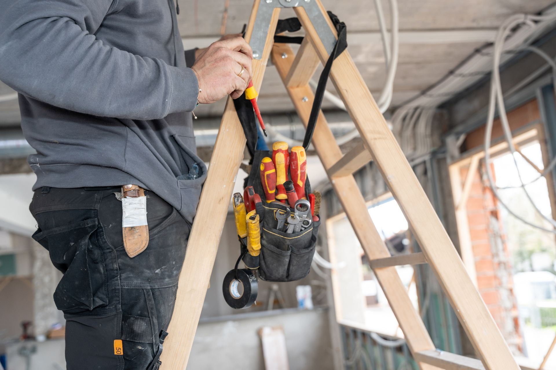 A construction worker stands by a wooden ladder holding a hanging tool pouch filled with screwdrivers and tape.
