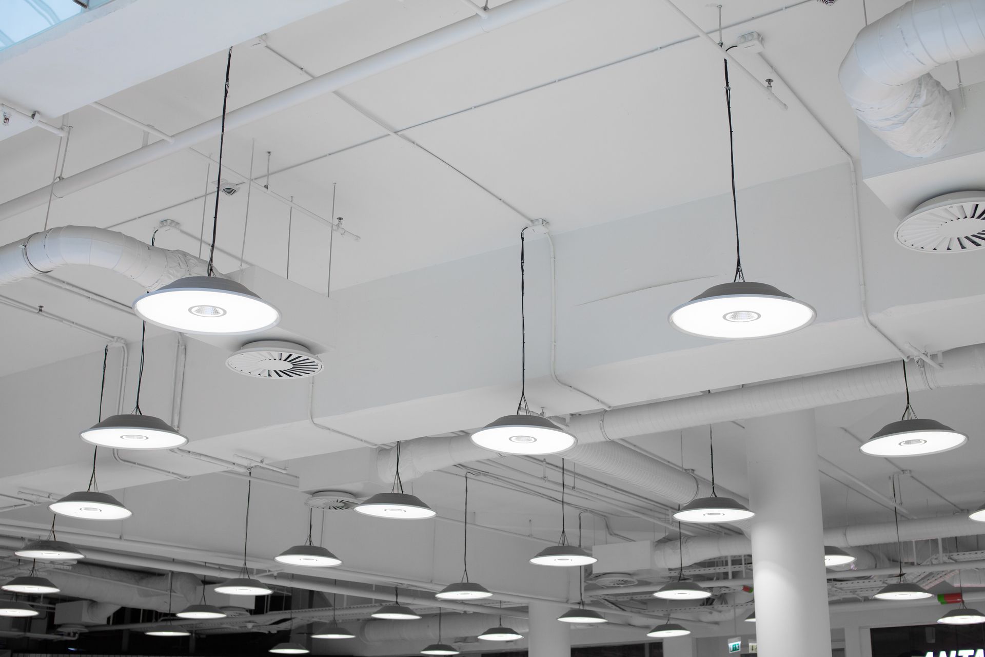A low-angle view of a white industrial ceiling featuring numerous hanging pendant lights and exposed ventilation ducts.