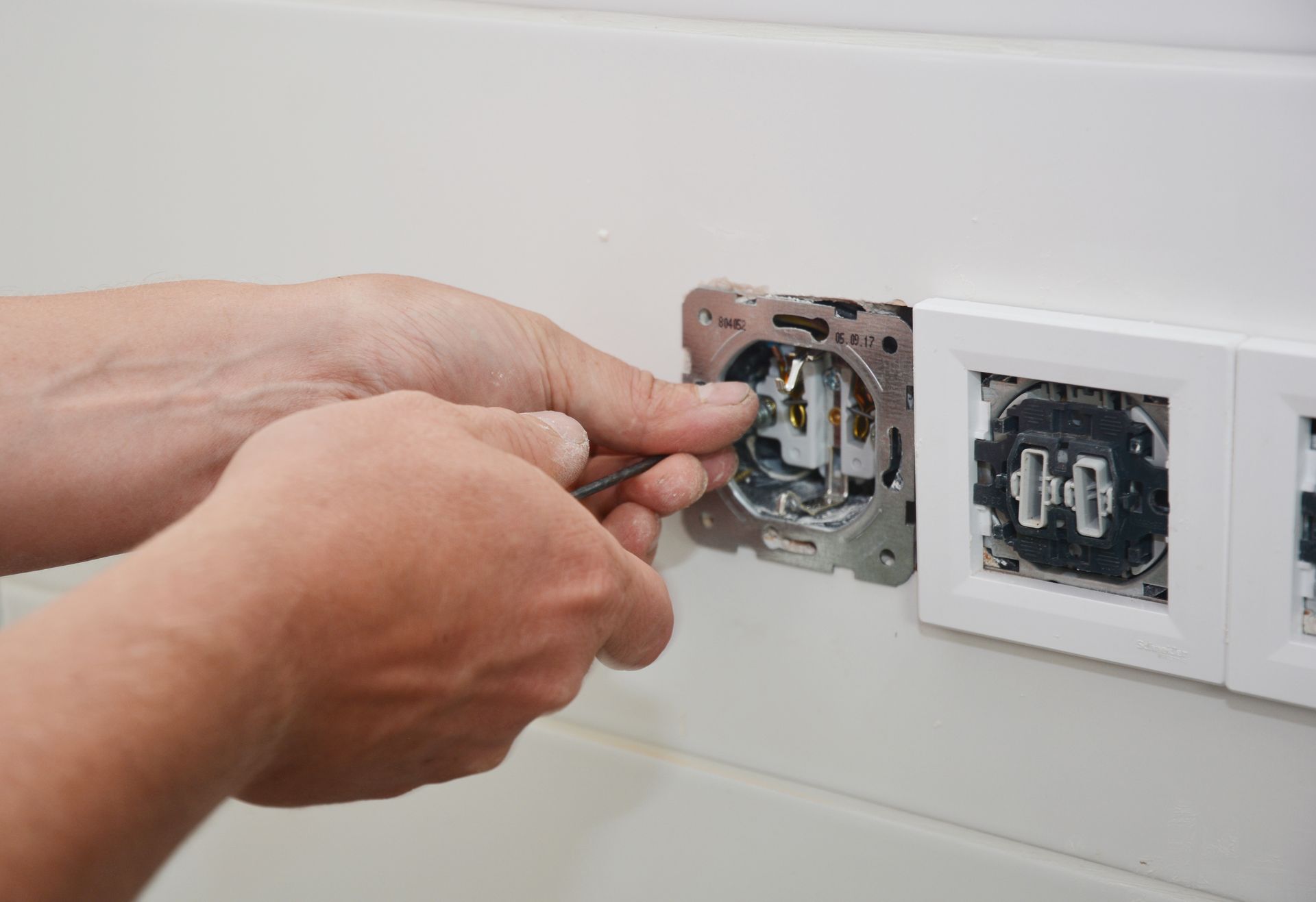 Hands using a screwdriver to install an electrical wall outlet in a white tiled wall.