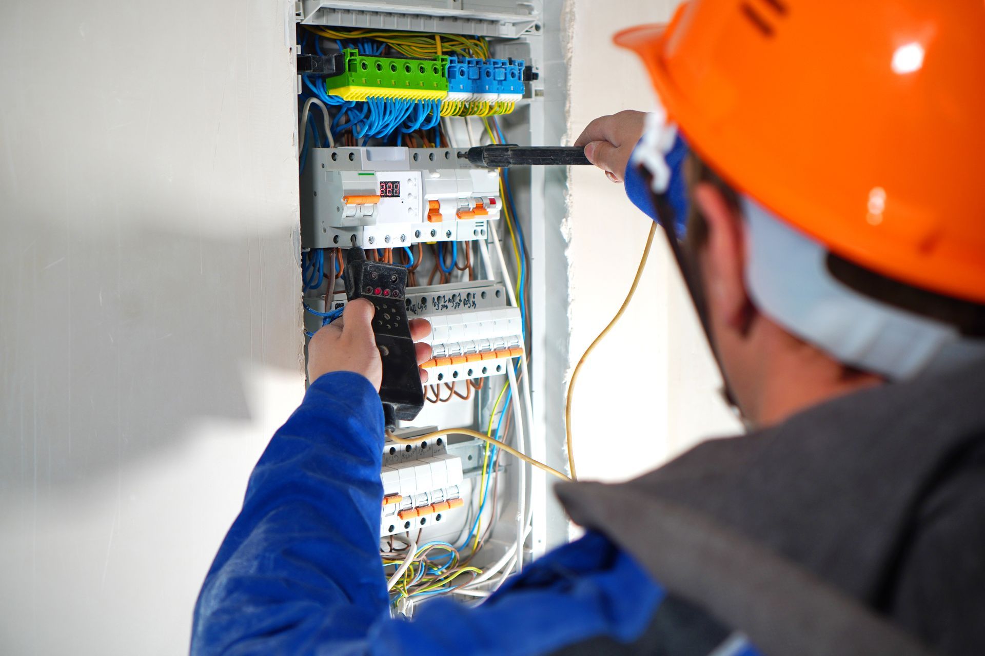 Electrician wiring a circuit breaker panel with a screwdriver, wearing an orange hard hat and blue jacket.