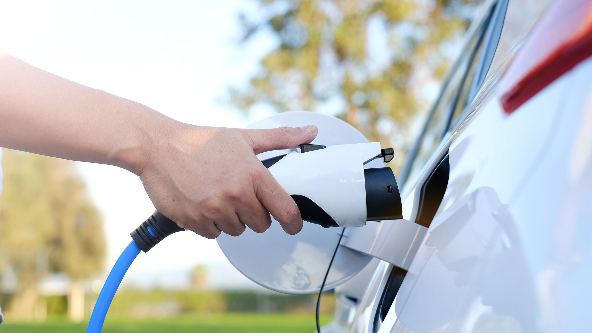 A hand plugging a blue electric vehicle charging cable into a white car in an outdoor setting.