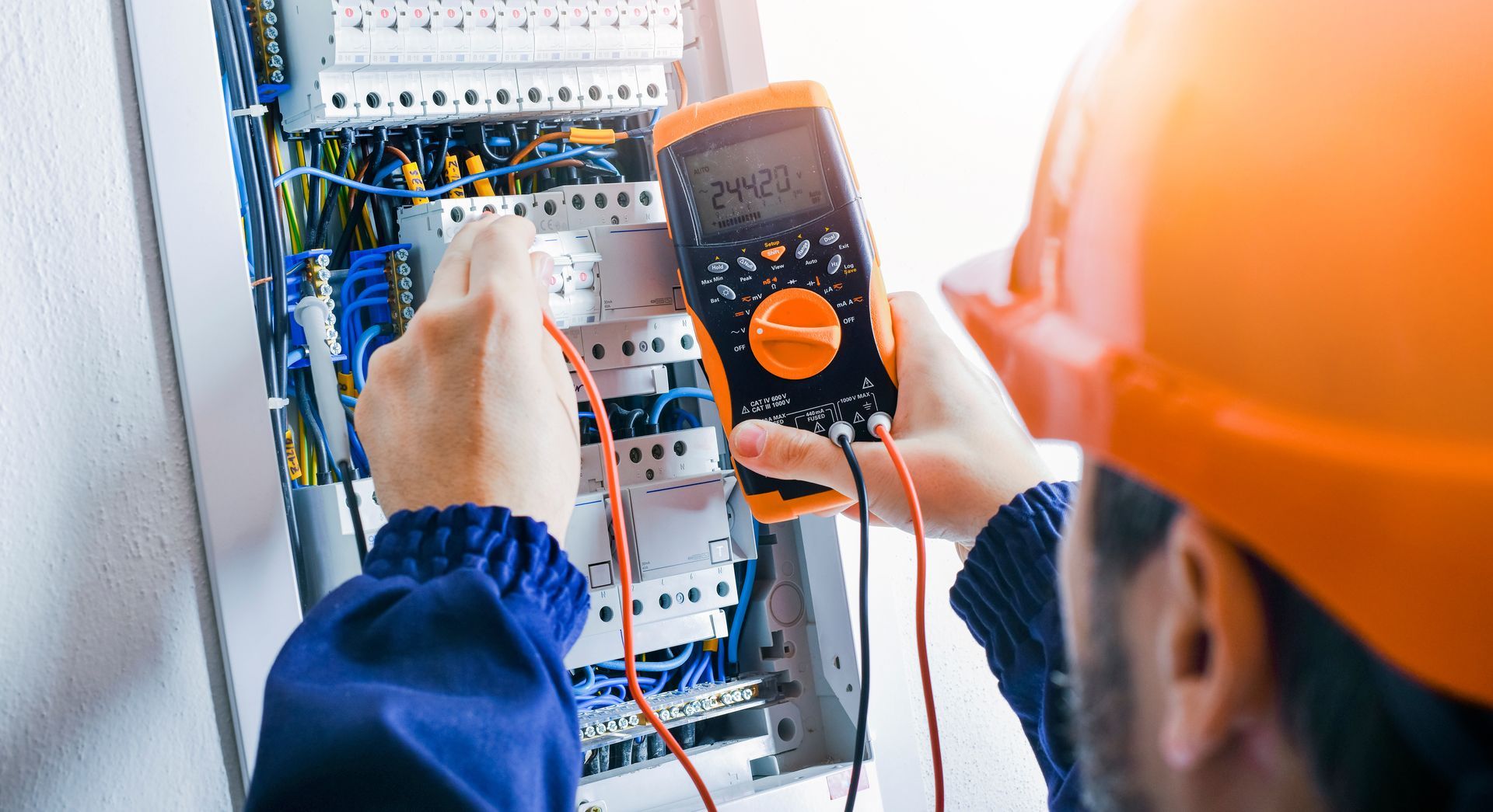 A professional in a hard hat uses a multimeter to test a circuit breaker panel.