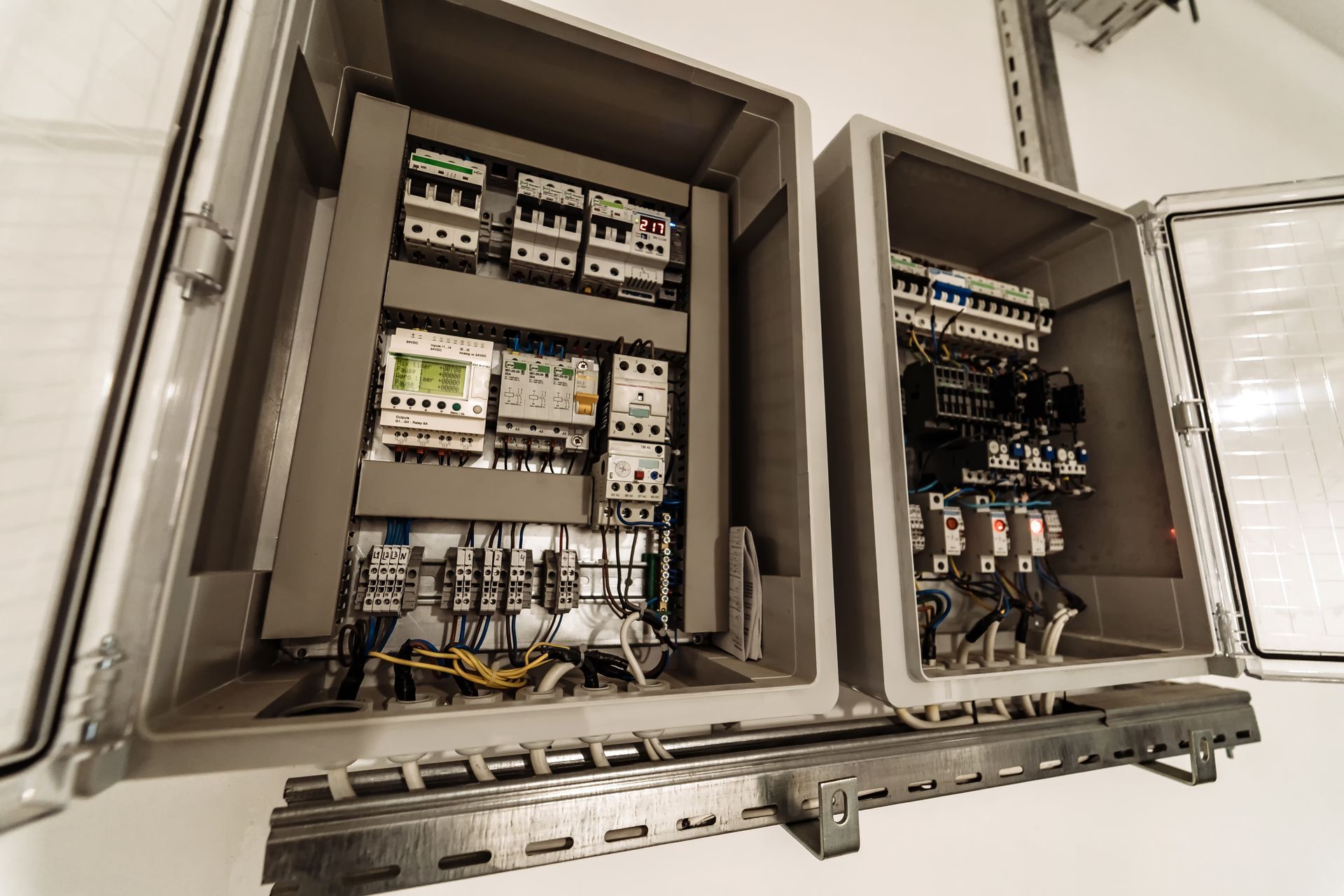 Two open gray electrical control panels mounted on a white wall, featuring internal circuit breakers and wiring.