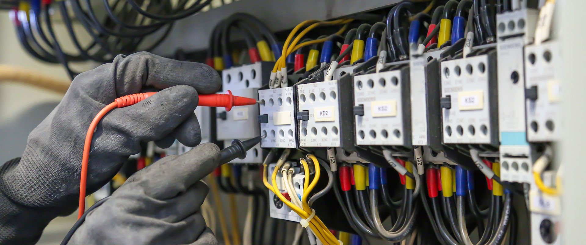 Gloved hands testing a wire in an electrical control panel with rows of breakers and cables