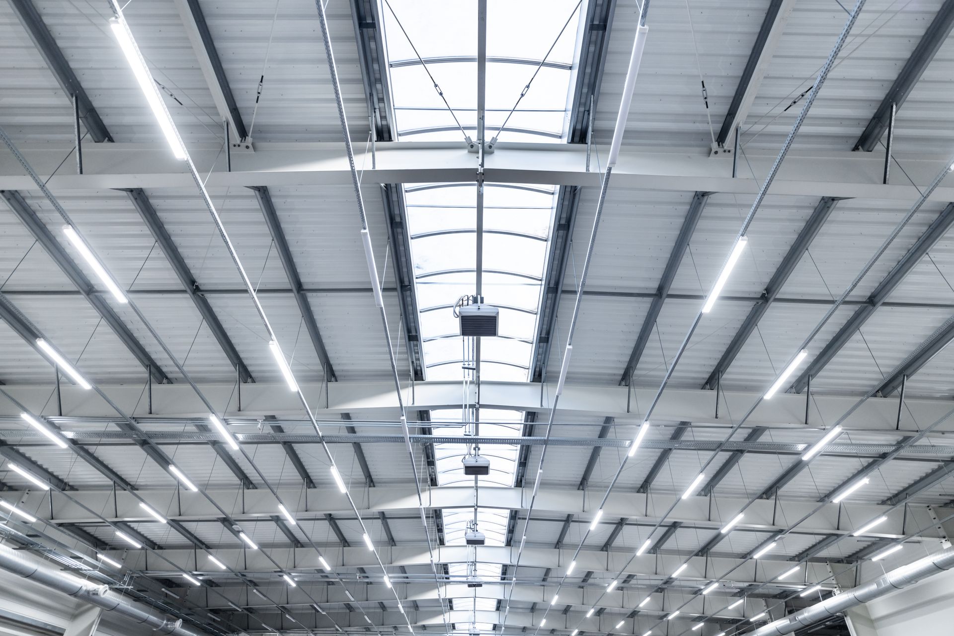 The high, industrial ceiling of a warehouse features white steel beams, parallel fluorescent lights, and a central skylight.