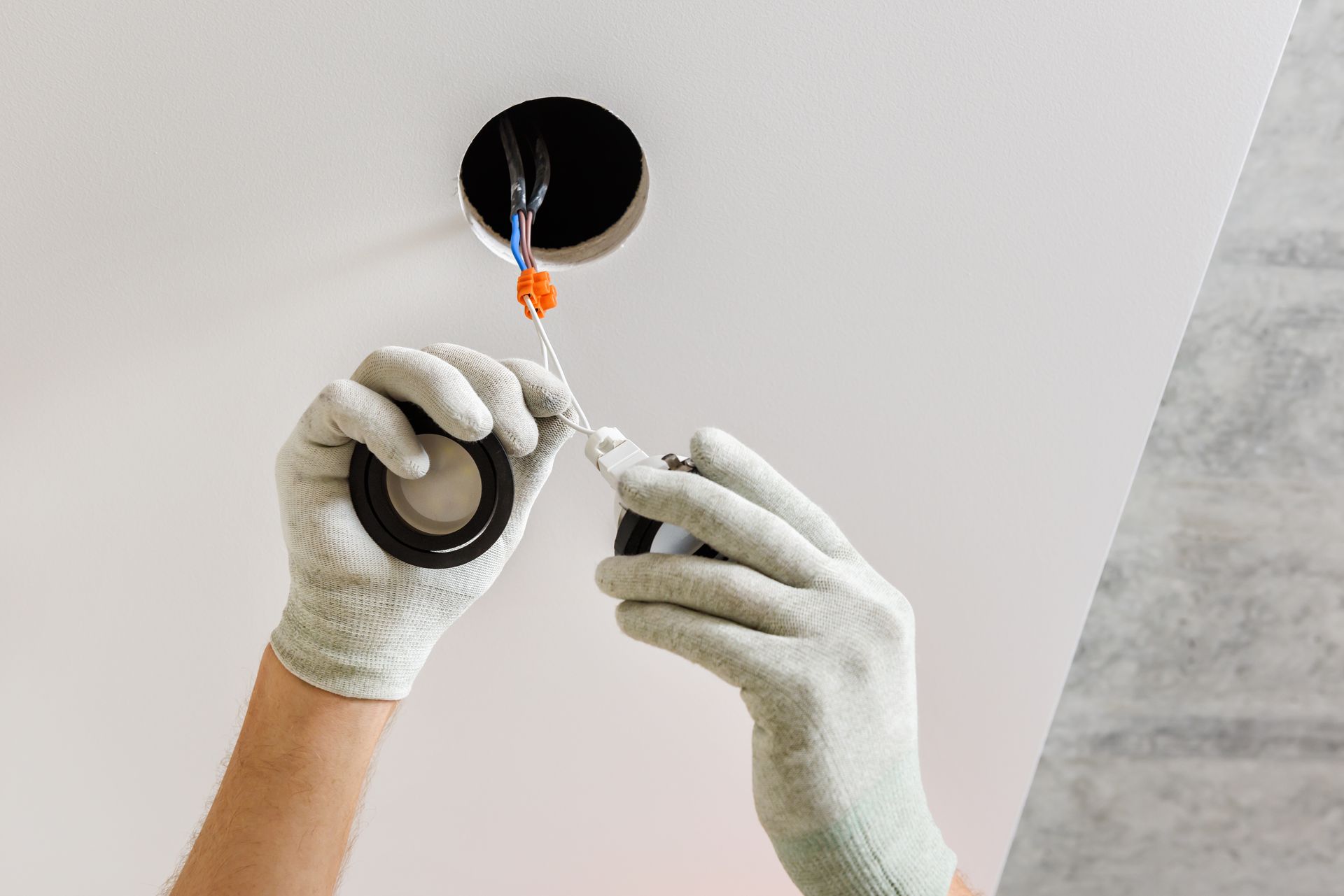 A pair of hands in light gray work gloves applying electrical tape to wires hanging from a hole in a white ceiling.