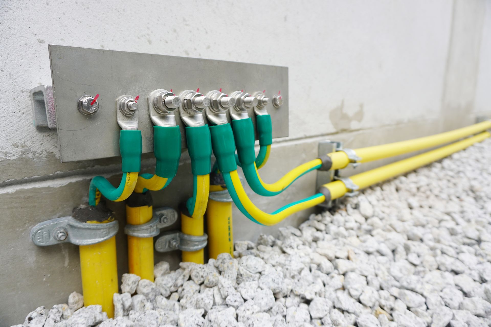 Yellow electrical conduits and green grounding wires connected to a metal junction box on gravel.
