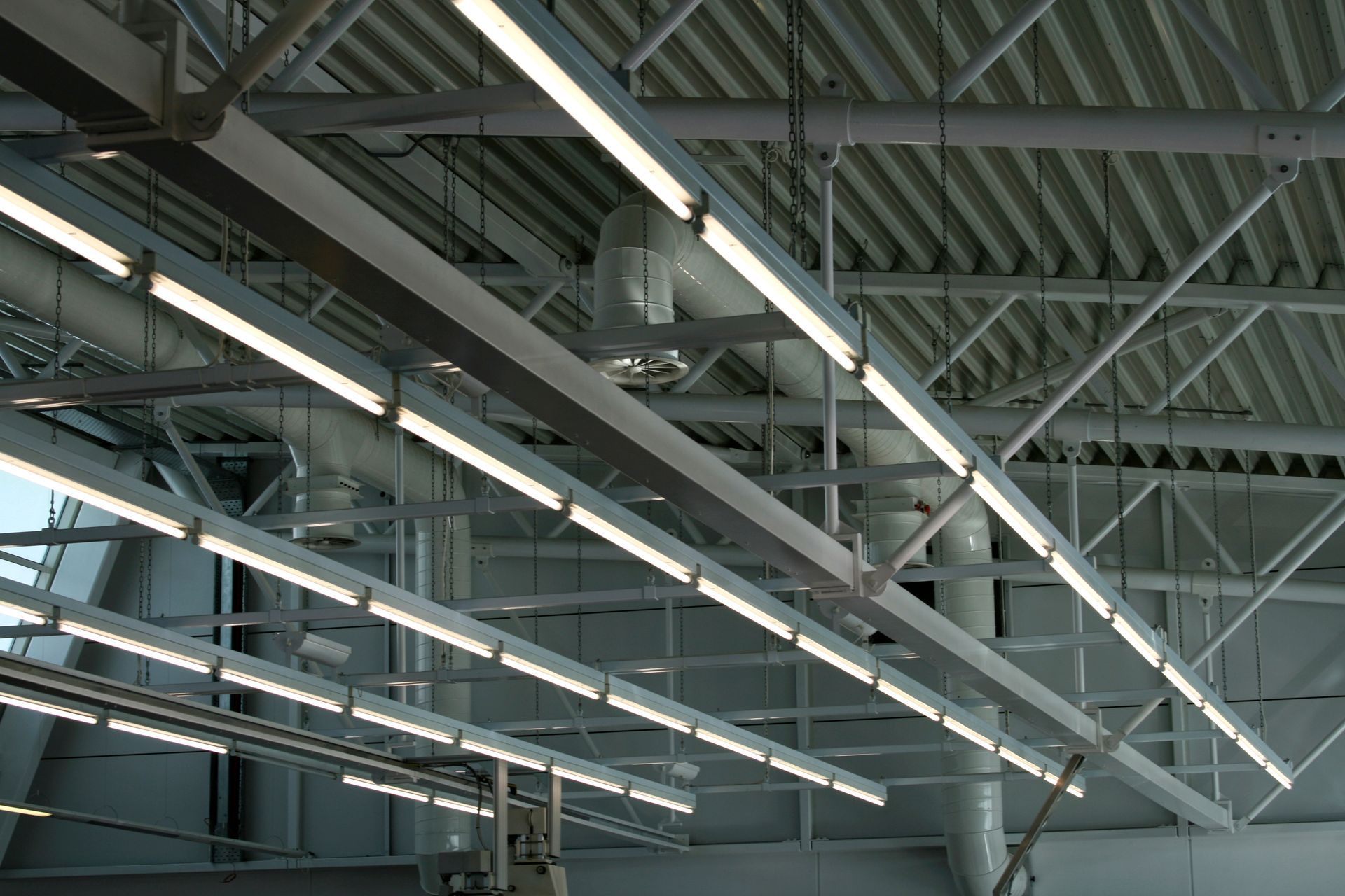 Long industrial linear light fixtures suspended from a white metal ceiling structure.