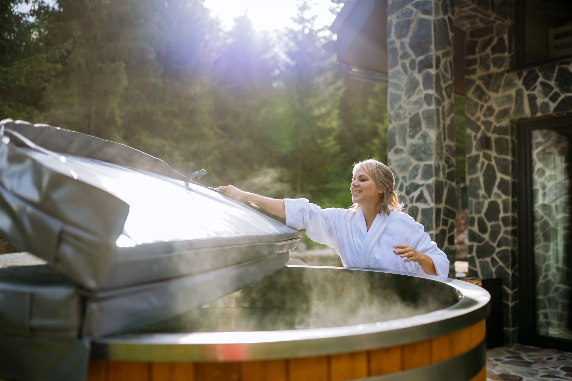 Woman in white robe opening a hot tub cover beside a stone building in a forest setting