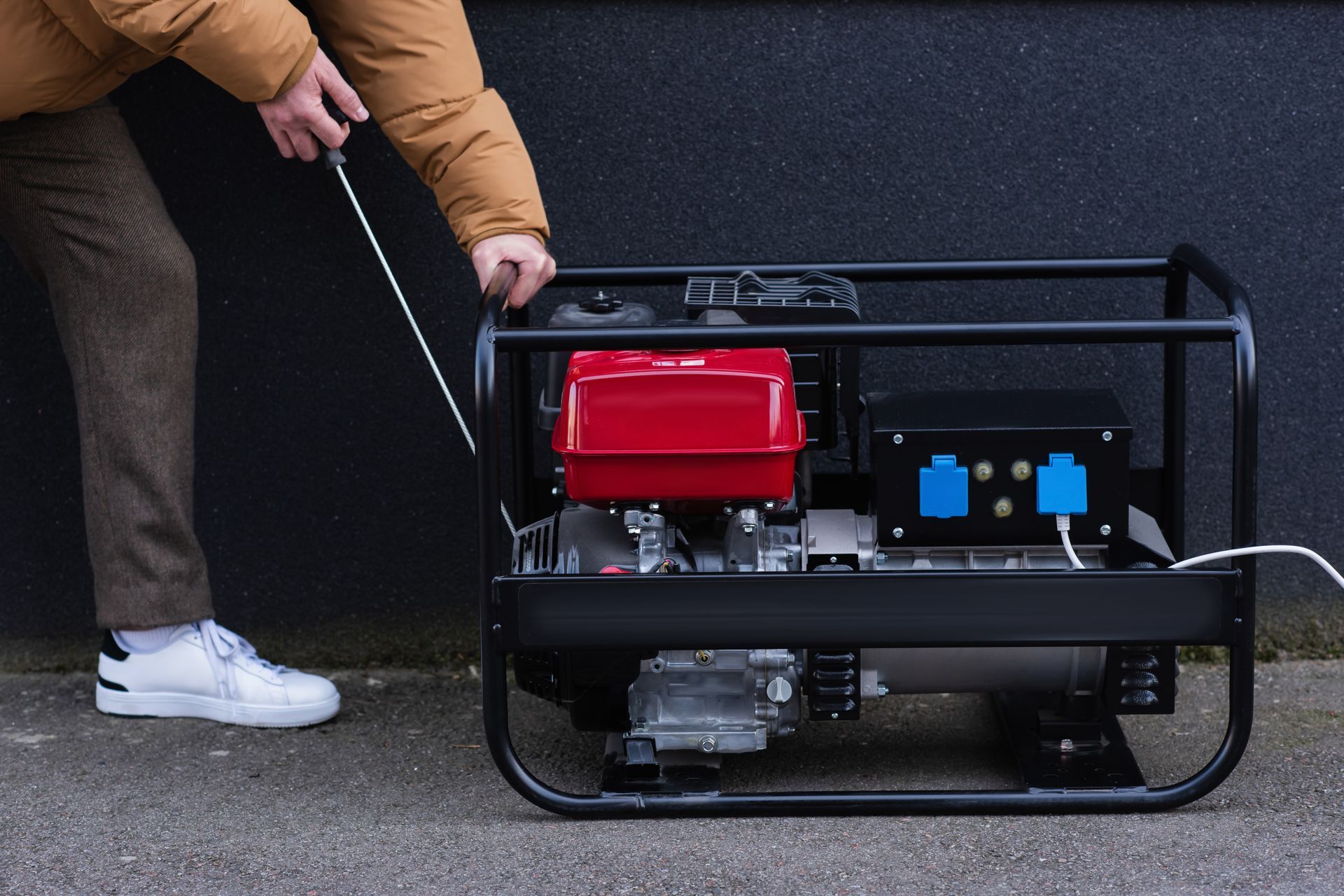 A person pulls the recoil starter handle on a portable, black-framed gasoline generator against a dark wall.
