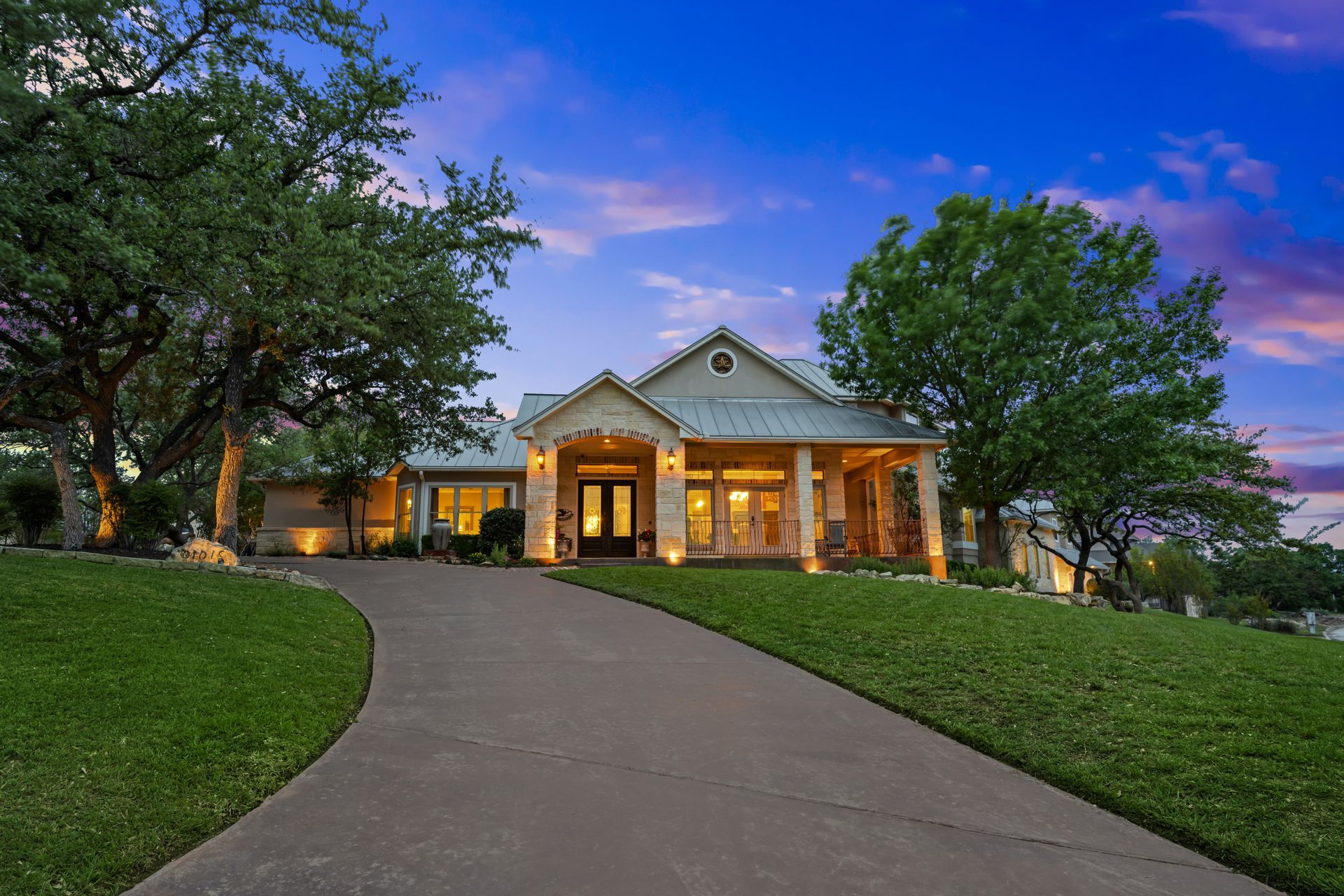 Grand house at dusk with warm lights, stone columns, and a curved driveway under a purple sky