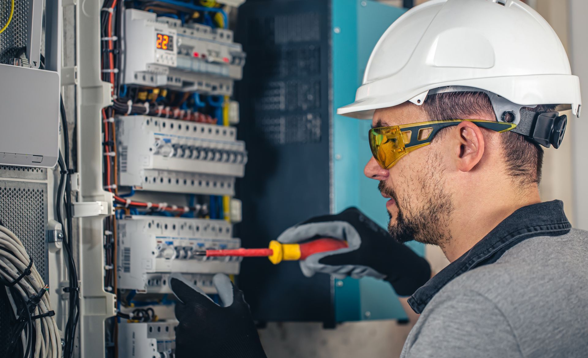 An electrician wearing a white hard hat and yellow safety glasses uses a screwdriver on a residential circuit breaker.