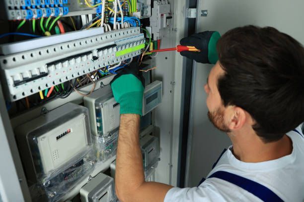 A technician in work clothes and green gloves uses a screwdriver to service an electrical panel.