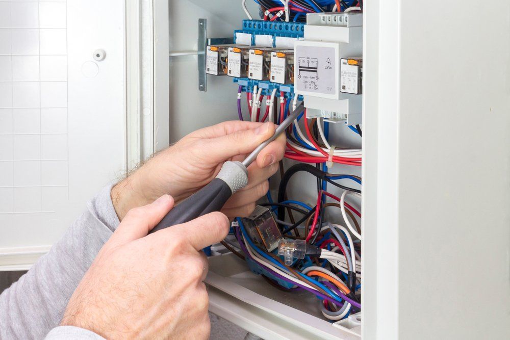 Hands using a screwdriver to work inside an open electrical control panel with wires and breakers.