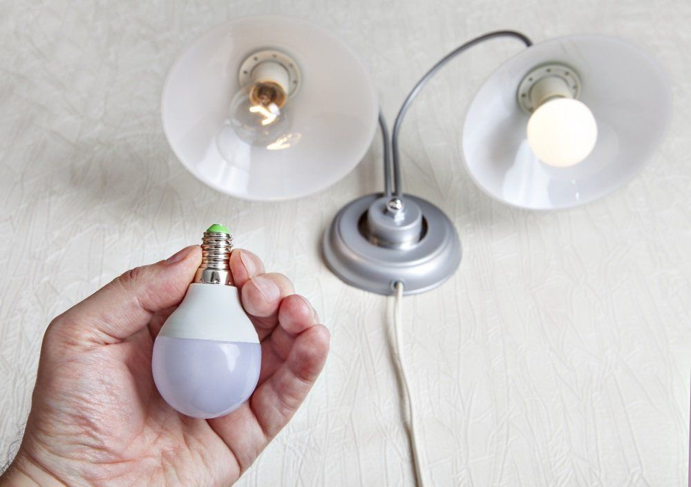 A hand holds a new LED lightbulb in front of a double-arm lamp with one clear and one frosted bulb installed.