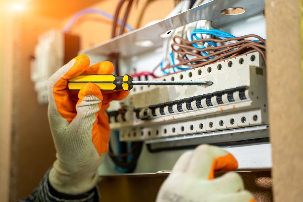 A person wearing safety gloves uses a screwdriver to work on a residential electrical circuit breaker panel.
