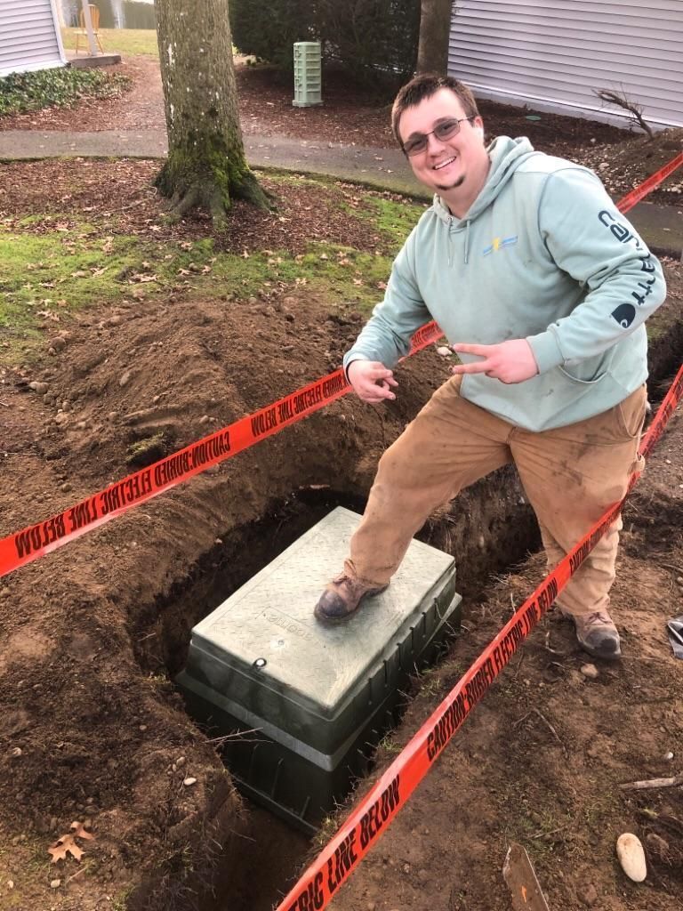 Man in a green jacket standing by an open utility box, smiling and pointing inside, near a taped-off trench.