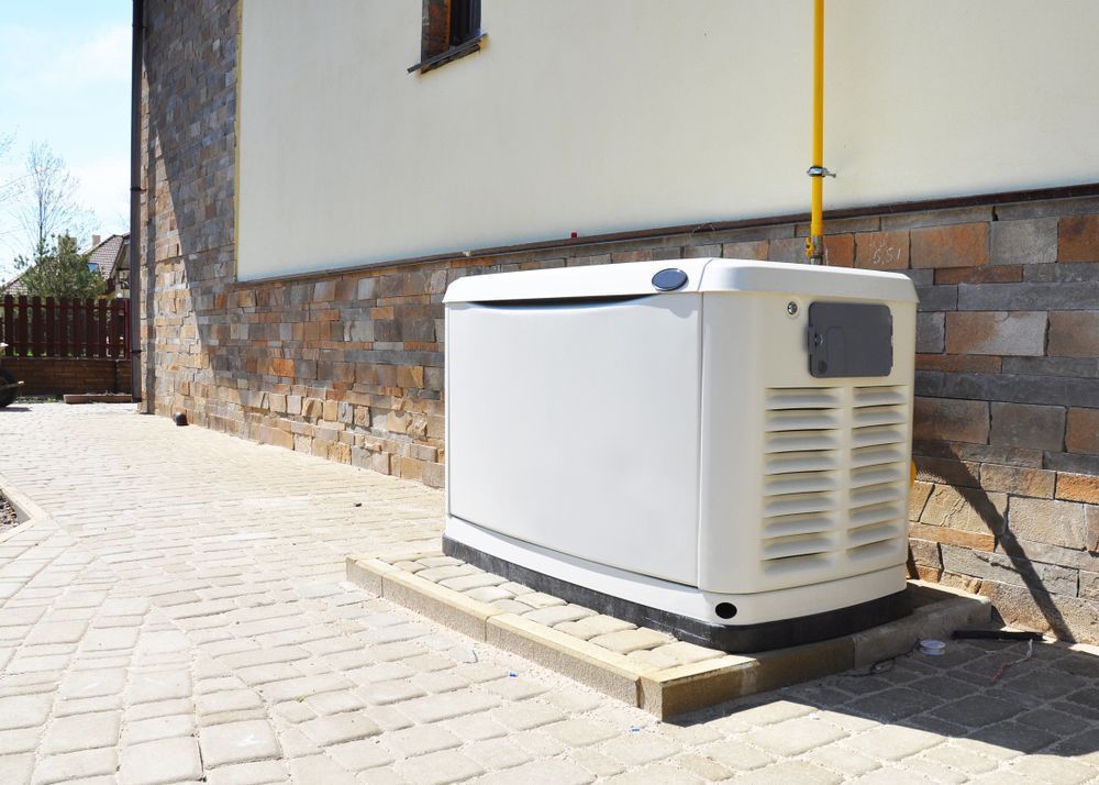 A white, rectangular standby home generator installed on a paved outdoor area next to a stone-walled house.