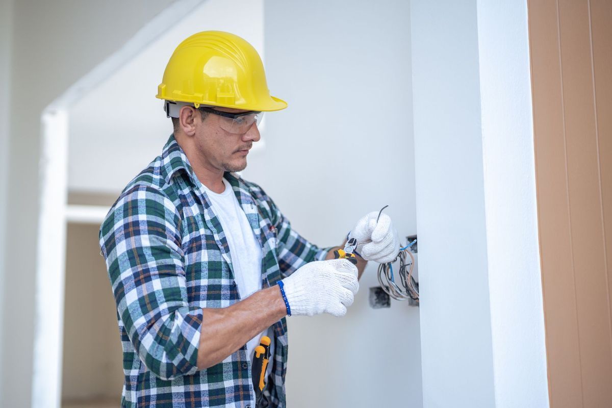 An electrician in a yellow hard hat and white gloves installs wiring into a wall outlet in an indoor setting.