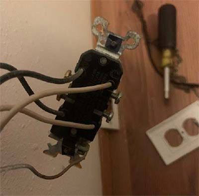 Close-up of a black electrical switch with wires, mounted on a wooden wall beside an outlet plate and screwdriver.