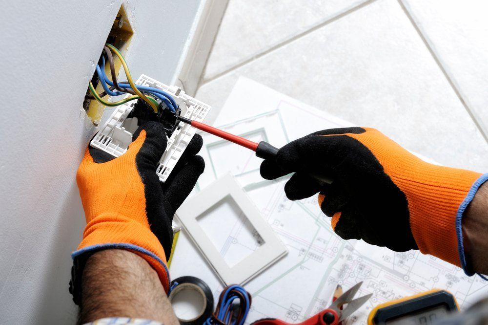 A person wearing orange and black work gloves uses a red-handled screwdriver to install a wall light switch.