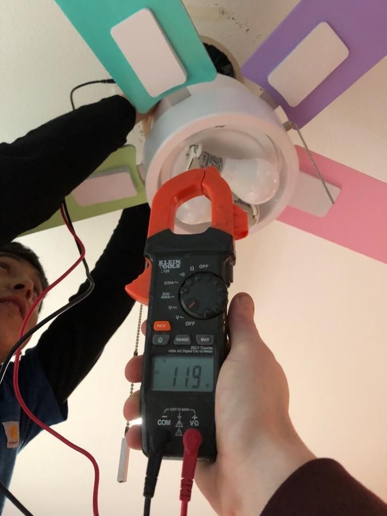 A person tests electrical voltage on a ceiling fan with colorful blades using a digital multimeter showing 119 volts.