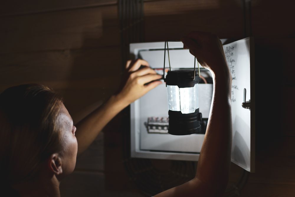 A person holds a battery-operated lantern to illuminate an open electrical breaker box in a dimly lit room.