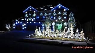 A house at night covered in blue and white Christmas lights, with a row of illuminated evergreen trees in the yard.